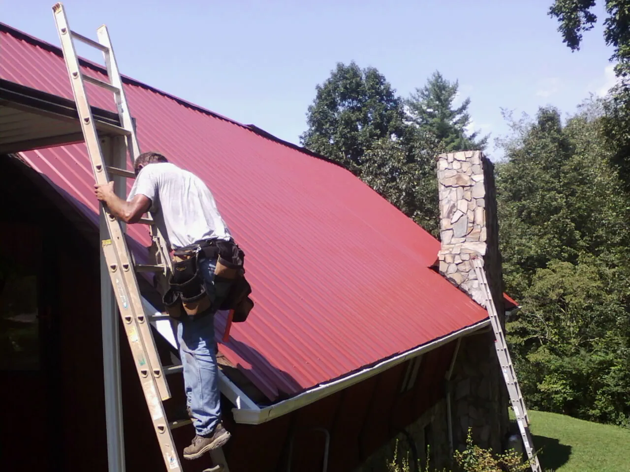 Roofer on a ladder beside a red metal roof and stone chimney — Soffit & Fascia Repair by Roofing Brothers in Collegedale